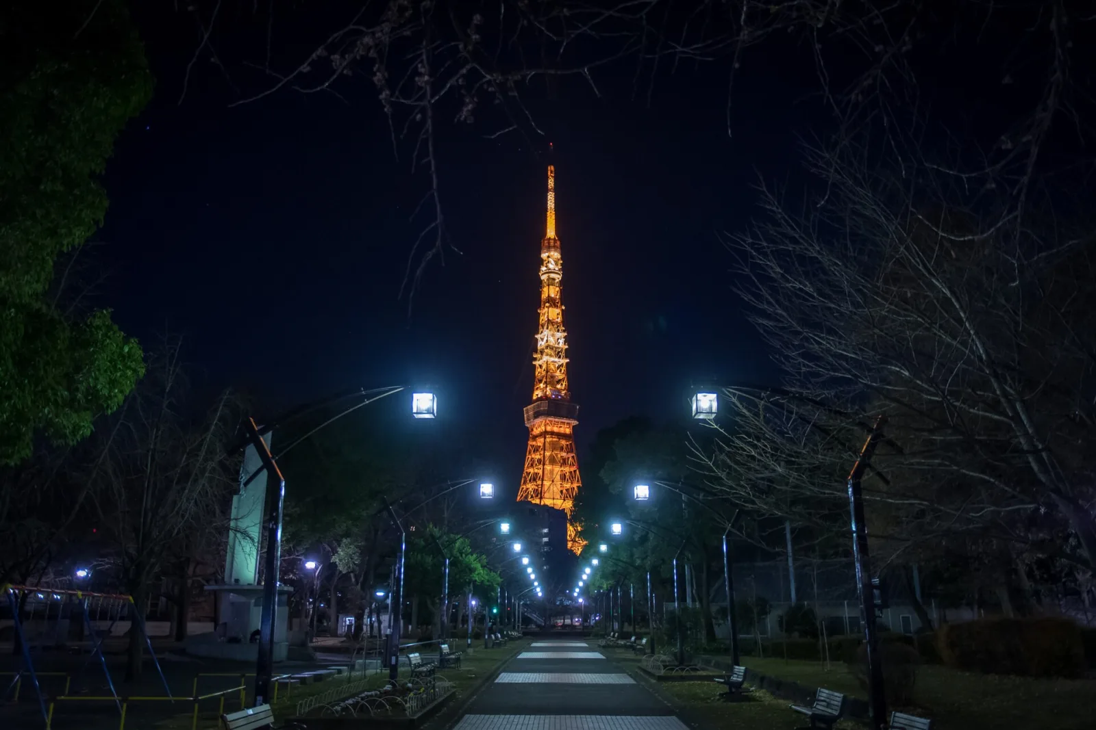 Tokyo Tower lit orange at night, seen from the lamplit path of Shiba Park